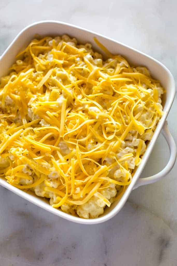 A square white baking dish with mac and cheese and shredded cheese topping, ready to go in the oven.